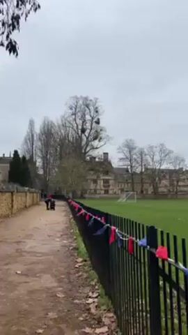 The video shows a dirt path alongside a black metal fence decorated with red and blue pennants. To the right of the fence is a green field with a soccer goal. Beyond the field, there are stone buildings. In the distance, two people, one pushing what appears to be a baby stroller, are walking along the path. The sky is overcast. Bare trees are scattered throughout the scene.