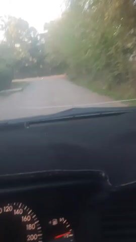 This is a first-person perspective video from inside a car driving on a rural, unpaved road. The dashboard shows a speedometer with the needle indicating a speed between 40 and 60. The windshield shows a dusty road curving to the left, flanked by trees and vegetation on both sides. The sun appears to be low in the sky, casting a warm light. The video is short and captures the continuous motion of the car.