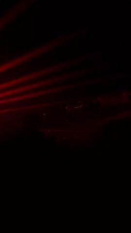 A dark video starts with complete blackness. Suddenly, spotlights are activated. Red lights beam from the ceiling of what appears to be a venue or club, with thin blue lights along the ceiling’s beams. The silhouettes of people can be seen standing in the foreground, facing the source of the light. The sound of music with a repetitive bass beat plays.