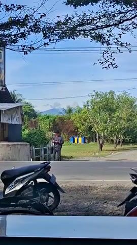 The video shows a street scene with several motorcycles parked in the foreground. In the background, a person stands near a building, looking at their phone. A white van drives past, followed by a red scooter with a rider. The sky is clear and blue, and there are trees and mountains in the distance. The scene appears to be a sunny day.