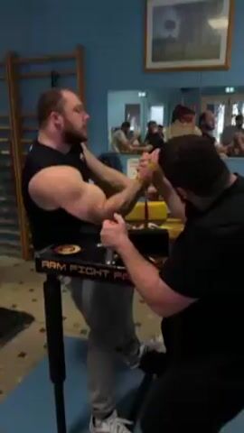 Two men engage in an arm-wrestling match on a table labeled "Arm Fight Pad." The man on the left, wearing a black tank top and gray pants, has a beard and tattoos on his arm. He is seated and uses his left hand to grip the table. The man on the right, wearing a black shirt, is also seated and appears to be controlling the arm-wrestling match. A yellow strap secures their wrists together. The room is painted blue with a checkered floor. A ladder leans against the wall on the left side. A mirror on the back wall reflects the arm-wrestling match and other individuals watching. A framed picture hangs above the mirror.