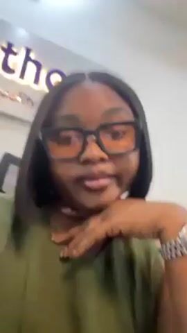 A woman with dark, shoulder-length hair, wearing a green blouse and black-framed glasses with light-colored lenses, is looking at the camera. Her left hand is resting on her face. She has a silver watch on her left wrist. A sign with the word "ortho" in purple lettering is visible in the background. The setting appears to be indoors, possibly an office.