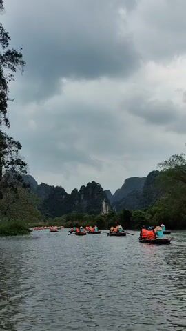 The video shows a river scene with many small boats, each carrying several people wearing orange life vests. The boats are spread out across the river, with some clustered together and others more isolated. People are paddling the boats. The river is surrounded by green vegetation and dramatic, rocky hills, which are visible in the background. The sky is overcast with gray clouds.