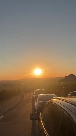 A sunset is shown in this video. The sun is setting over a landscape of vineyards. There are cars parked on a road overlooking the vineyards. There are also a few people walking or standing near the cars. The sun is bright and the sky is orange and yellow. The video zooms in on the sun, showing it partially eclipsed by the moon.