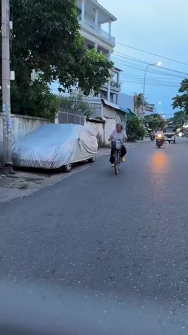 A wide shot shows an elderly woman riding a bicycle down a paved street. She is wearing a white and blue patterned shirt, black pants, and yellow shoes. The woman has gray hair pulled back. She is facing left as she rides the bicycle, which has a basket on the front. In the background, a car is covered with a silver tarp. A motorcycle with a rider is visible further down the street, its headlight illuminated. Trees and buildings are on the left and right sides of the street. The sky appears to be dusk.