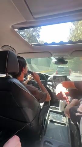 The video shows a group of friends traveling in a car on a sunny day. The driver, a young man with dark hair and a brown shirt, is holding a microphone and appears to be singing or rapping. The front seat passenger, who has curly brown hair and is wearing a gray shirt, is also singing along. A third person is partially visible in the front seat and is holding a water bottle. A fourth person in the back seat is holding a portable speaker with colorful dials and buttons. The car has a sunroof, and the road ahead is visible with other cars. The interior of the car is black, and the center console holds a phone. The atmosphere in the car appears to be lively and fun.