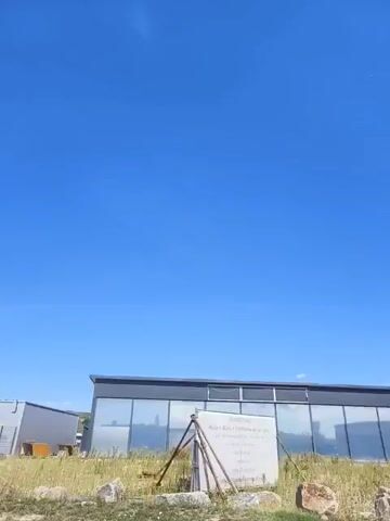 The video captures a panoramic view of a sunny day with a clear blue sky. It pans across various buildings, some of which appear to be industrial or commercial. Prominently featured is a large solar panel array on the left side of the screen. As the camera moves, different structures come into view, including buildings with large windows and signage. One building displays the name "pucher Wärme- und Wassertechnik GmbH" along with a yellow faucet logo. Another building has signage for "EP-Zierlinger" and a website address. In the foreground, there is some vegetation and fencing. The overall impression is an outdoor scene in a developed area, possibly a business park or industrial zone. There are no people visible in the video.