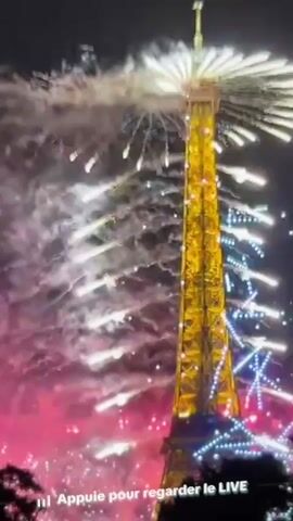 This is a video of the Eiffel Tower at night with fireworks exploding around it. The fireworks are colorful, with shades of pink, purple, and white. The Eiffel Tower is illuminated and decorated with lights. There are also some trees in the foreground. Text at the bottom of the screen says "Appuie pour regarder le LIVE," which translates to "Press to watch LIVE."