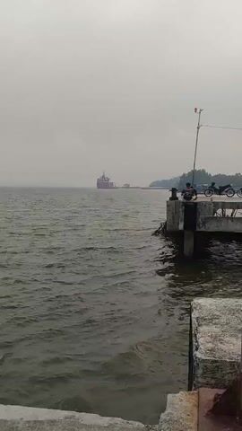The video shows a body of water with choppy waves. On the right side of the screen, there is a concrete pier. Two people are sitting on the pier. A motorcycle is parked behind them. In the distance, a large red and white ship is visible on the horizon. The sky is overcast and the overall atmosphere appears hazy.