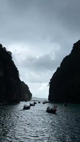 A drone shot captures a body of water between two large, dark, rocky cliffs. A fleet of small, dark boats with people on them navigates the water, moving away from the camera and towards a bright horizon obscured by heavy, gray clouds. The water is choppy with moderate waves, and the overall lighting suggests an overcast day.