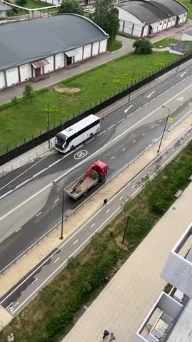 The video shows an aerial view of a road and sidewalk. A white bus is traveling on the road. A red truck with a crane is parked on the road next to a sidewalk. A person is walking on the sidewalk, away from the truck. The sidewalk is bordered by a fence and grass. In the foreground, there is a building with balconies. The weather appears overcast.