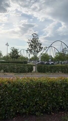 The video shows a view of an outdoor area with a roller coaster in the background. In the foreground, there are hedges and trees. There is a billboard with text that says "BIENVENUE A MONACO" and an image of a cartoon character. There are cars parked in the background. The sky is cloudy. The video pans slightly to the right.
