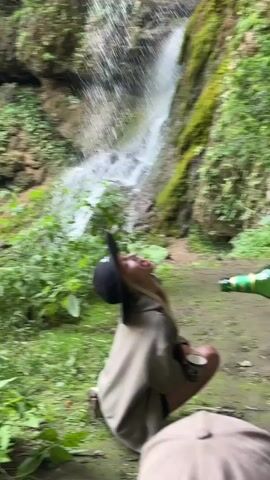 A young woman wearing a dark baseball cap and a light-colored hooded jacket kneels in a natural setting with lush green foliage and a waterfall in the background. She is looking upwards with her mouth open and tongue extended, as if to catch water from the waterfall. A green bottle is visible in the right side of the frame, held by an unseen person. The woman holds a small white cup in her left hand.