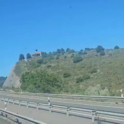 The video shows a low-angle shot of a small building with a tower and a cross on top, resembling a chapel or shrine, situated on a grassy hill against a clear blue sky. The hill is covered with sparse vegetation, including bushes and some trees. A road with a guardrail is visible in the foreground, suggesting the video was taken from a moving vehicle. The camera zooms in slightly on the building.