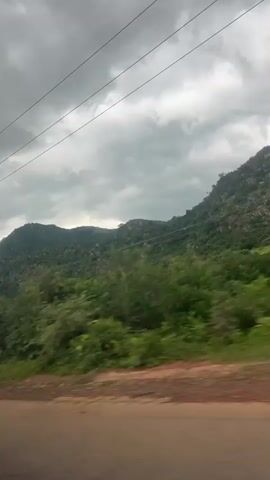 This is a POV video captured from a moving vehicle. The camera is tilted slightly downwards, showing a dirt road in the foreground and lush green trees and hills in the background. Power lines are visible stretching across the grey, cloudy sky. The vegetation is dense, and the terrain appears hilly and rugged. There are no people or animals visible in the frame. The lighting suggests an overcast day, possibly before or after rain. The motion blur indicates the vehicle is in transit.