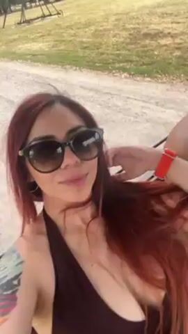 A woman with long red hair and sunglasses takes a selfie video while riding in a golf cart. She is wearing a dark bathing suit top and has a tattoo on her arm. A man without a shirt is sitting next to her and another man is driving the cart. They are driving on a dirt path surrounded by trees and grass. The weather appears to be sunny.