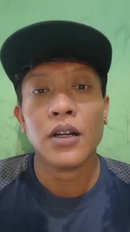 A close-up, head-and-shoulders shot of a person wearing a black baseball cap and a dark t-shirt. The person has brown skin and short, dark hair. They are looking directly at the camera and speaking. The background is a solid green wall. The video is shaky, suggesting it was recorded handheld.