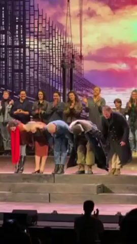 The video shows a group of people on a stage taking a bow at what appears to be the end of a performance. The background features a painted backdrop with an abstract, possibly urban landscape. The performers are dressed in various costumes, suggesting a theatrical production. In the foreground, the audience is visible in silhouette. The performers bow and then stand upright, holding hands. The group appears to be acknowledging applause.