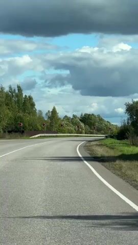 This is a first-person view of a car driving on a winding asphalt road. The road has white lane markings and is bordered by grass and trees. There are several red and white directional signs indicating curves in the road. The sky is partly cloudy with large, dark gray clouds interspersed with patches of blue sky. The sun casts shadows on the road, suggesting it is daytime.