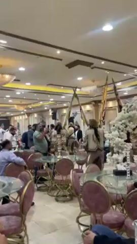 Inside a brightly lit banquet hall, a group of men in traditional Middle Eastern attire are parading through the room, carrying tall, decorative flags. They weave between tables set for a reception, some occupied by seated guests watching and recording the spectacle with their phones. The room is decorated with white floral arrangements and reflective surfaces, adding to the celebratory atmosphere. Other guests and videographers capture the event as it unfolds.