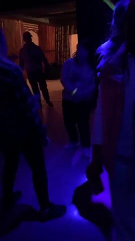 The video shows a dimly lit room with people dancing. The lighting is a mix of purple and red. In the foreground, a person is partially visible from the shoulders down, wearing dark clothing. To their right, another person in a light-colored top and dark pants is dancing. Further in the background, several other individuals are visible, some dancing and others standing. One person in the background, wearing a light blue shirt, appears to be dancing. Another person in a grey hoodie and blue jeans is standing still. In the middle of the frame, a person in a grey top and black pants is dancing enthusiastically, with their arms outstretched. The floor appears to be tiled. There are dark, textured walls or partitions in the background, and a colorful mural or decoration is visible on the upper right wall. A person in the middle ground appears to have fallen to the floor while dancing. The scene suggests a party or club environment.