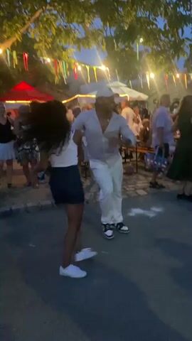 A group of people are dancing outdoors in the evening. A man and a woman are dancing in the foreground, holding hands. The man is wearing a white shirt, white pants, and a white hat. The woman is wearing a white top and a dark skirt. In the background, other people are visible, some dancing and some sitting at tables. String lights are hung overhead, and there are trees and buildings in the background. The overall atmosphere appears festive.