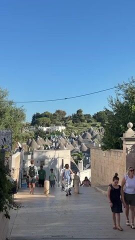 The video pans across a sunny, outdoor scene in a European town. In the foreground, a paved pathway slopes downward, with people walking and sitting. On the right, a young woman in a black dress walks towards the camera. To the left, a group of people are walking down the path, one carrying a backpack. In the background, a cluster of conical, white-roofed buildings, characteristic of Alberobello, Italy, are visible on a hillside under a clear blue sky. Further to the left, a white church with a cross on its roof is seen, with a fountain in front of it. A street lamp with lights and a security camera is visible mounted on a pole. Olive trees and other greenery are scattered throughout the scene.