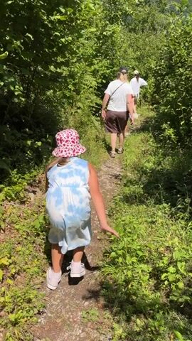 The video begins with a shot from behind of a young girl in a tie-dye dress and a pink hat, walking up a dirt path in a wooded area. She is followed by two adults, one in a white shirt and shorts, and another person partially visible in a red shirt. The path is surrounded by lush green foliage. The camera then transitions to a selfie shot of a blonde woman with sunglasses and a white dress, smiling at the camera while standing in the same wooded environment. The video appears to capture a casual outdoor excursion.