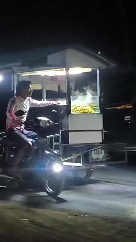 A person is sitting on a motorcycle that is pulling a food cart. The food cart has a canopy with lights and steam is rising from inside, indicating food is being cooked. The person on the motorcycle is wearing a long-sleeved shirt and shorts and is reaching out with their right hand. The motorcycle has its headlight on. The scene appears to be at night, with the area around the cart and motorcycle illuminated by artificial lights. The ground appears to be unpaved.