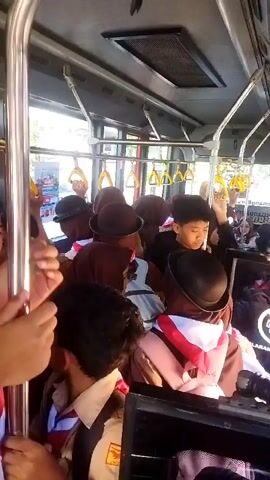 The video shows the interior of a crowded bus. Many young people, likely students, are standing and holding onto overhead bars. They are wearing uniforms that include brown shirts, red and white sashes, and hats. Some of them are looking around or at their phones. The bus appears to be in motion, as evidenced by the natural lighting that shifts. The environment is utilitarian, with metal poles, plastic seats visible in the background, and ceiling vents. The overall atmosphere suggests a busy commute or field trip.