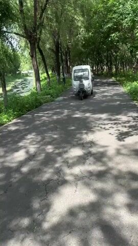 The video shows a person driving a white three-wheeled motorized vehicle on a paved path in a park. The vehicle is carrying cargo in its rear bed. The path is lined with trees on both sides and a body of water is visible to the left of the path. The video then pans to show another person in the vehicle, adjusting their red sunglasses. The scene appears to be a sunny day.