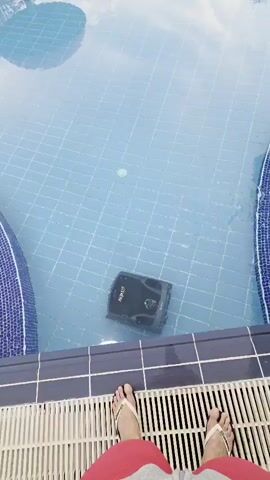 A robotic pool cleaner is shown floating in a swimming pool. The pool is tiled with blue and white tiles. The water is clear and blue, reflecting the sky and surrounding trees. The perspective of the shot is from the edge of the pool, looking down at the cleaner. The feet of a person wearing flip-flops are visible in the foreground. The robot appears to be black with a green light on its top.