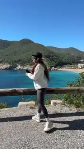 A person wearing a baseball cap, sunglasses, and a sweatshirt with "BROOKLYN" on it is standing on an overlook with a picturesque beach in the background. The beach is a sandy cove with crystal-clear turquoise water. The surrounding landscape features green hills and some buildings. The sky is a clear blue. The person is holding something in their hand, possibly a phone or small object. The overlook has a wooden rail.