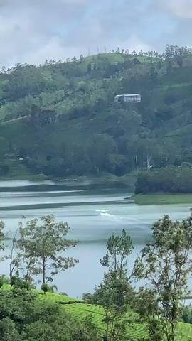 A white seaplane is seen taking off from a lake in a mountainous region. The lake is surrounded by lush green hills covered with trees and tea plantations. There is a building visible on the hillside in the background. The sky is cloudy.