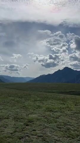 The video pans across a vast, open meadow with green grass and rolling hills. In the background, a range of majestic mountains stretches across the horizon, their peaks casting shadows under a partly cloudy sky. The clouds are a mix of white and gray, with some areas of sunlight breaking through. The landscape appears serene and untouched, evoking a sense of natural beauty and tranquility.