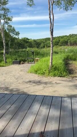 The video shows a view from a wooden deck looking out at a grassy area with trees and a blue sky. In the foreground, a pair of bare feet are crossed and appear to be resting on the deck. In the middle ground, there is a gravel area with a grill and a small wooden stool. The background consists of lush green trees and a bright blue sky with white clouds. The overall scene depicts a relaxed outdoor setting.
