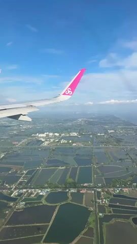 An aerial view from an airplane window shows a landscape of agricultural fields and bodies of water, likely aquaculture ponds, stretching towards a coastline. The airplane's wing, featuring a pink wingtip with a stylized logo, is visible in the upper left corner. The sky is blue with scattered white clouds. The scene is captured in daylight.