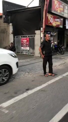 The media file shows a street scene in Sidoarjo, East Java. A man wearing glasses, a black vest over a black shirt, and black pants is standing on a paved sidewalk. He is looking to his left, with his right hand in his pocket and his left hand resting on his hip. He is wearing flip-flops.

To the left of the man, a white car is partially visible. Behind the man, there is a building with a dark awning and a metal fence. A sign on the fence reads "OPEN" in red and white lettering. Further back, there are more buildings and a motorcycle parked on the street.

The lighting suggests it is daytime, possibly late afternoon, as the light is soft and there are shadows. The weather appears to be clear. The scene is urban, with buildings and paved surfaces. There are no explicit cues about the man's emotions, but his stance is relaxed.

Visible text includes "OPEN" on a sign. There are also partial signs on the buildings in the background, including what appears to be "76" and "APEL" which are likely brand names or advertisements.