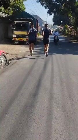 The video begins with a medium shot of two individuals running down a paved road from a rear perspective. To the left of the road, a yellow and black truck is parked. The runners are wearing dark-colored athletic shorts and t-shirts, with one runner in a blue shirt and the other in a black shirt. They are casting long shadows on the asphalt, indicating a low sun angle, likely morning or late afternoon. Beyond the truck, there are buildings and trees lining the road. In the distance, a person on a white motorcycle is visible. The camera then shifts to a close-up of a motorcycle's digital dashboard, displaying the number 22, presumably a speed reading, and reflecting the rider's hand. The perspective is now from the rider's viewpoint on the motorcycle, looking down at the dashboard.