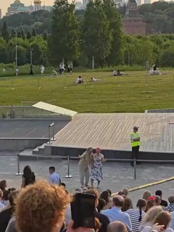 The video shows a performance in an outdoor amphitheater. Two people, a man and a woman, are standing center stage. The man is wearing a light-colored shirt and pants. The woman has pink hair and is wearing a floral dress. They are interacting with each other in a playful manner, with the man gesturing and the woman smiling. An audience is visible in the foreground. In the background, people are seated on a grassy slope, and a person in a yellow vest stands near a stage structure. The lighting suggests it is late afternoon or early evening.