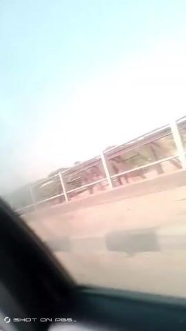 The video, shot from a moving vehicle, presents a view of a red metal bridge. The camera is positioned to the side, looking through the bridge's railing. Outside of the bridge structure, the landscape appears to be arid with some trees and vegetation. The sky is clear and pale blue. The vehicle is moving, blurring the view of the bridge and the surrounding scenery. The video is slightly shaky, and the camera angle is canted. "SHOT ON PES_" is stamped on the lower left corner of the frame.