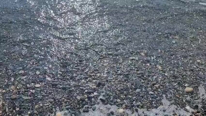 The video shows a close-up view of a pebble beach with small waves gently lapping at the shore. The pebbles are a mix of gray, brown, and white, and they are wet from the water. The water is clear and reflects the sunlight, creating a shimmering effect. The waves are small and appear to be coming from the right side of the frame, moving towards the left. There are no people or animals visible in the video. The overall impression is one of a calm and peaceful beach scene.