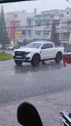 A white pickup truck drives through a street in the rain. Buildings line the street, some with signs in Vietnamese. Cars and motorcycles are also visible on the wet road. The sky is overcast, and rain is falling heavily. The scene appears to be an urban environment.