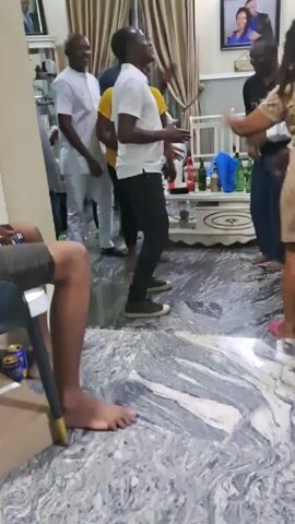Several people are gathered in what appears to be a home living room, dancing and socializing.  Most of the men are dressed casually, some in button-down shirts and slacks. A few are wearing t-shirts.  A woman in a patterned dress joins the group later in the video.  There are several bottles and cans on a table, suggesting they are enjoying beverages.  The room is decorated with framed pictures on the walls and a chandelier on the ceiling. The floor has a gray and white marbled pattern. One person is sitting with their legs visible, resting on a small chair.  The atmosphere seems festive.