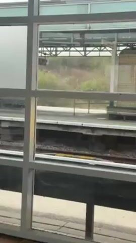 The video shows a train passing by a window. The window has a white frame and is divided into multiple panes. The train is moving quickly, making it difficult to discern details, but it appears to have white and red coloration and features the name "Avanti" displayed. The environment seen through the window is a train station platform and track.
