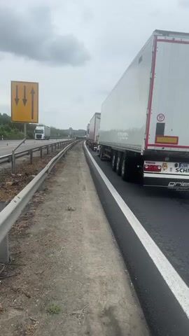 The video shows traffic stopped on a highway, likely due to congestion. Several large semi-trucks with trailers are lined up, along with a car and a caravan. A person is seen briefly exiting a blue SUV. The sky is overcast and the surrounding environment features green fields and trees. A road sign with yellow arrows pointing ahead is visible on the side of the road.