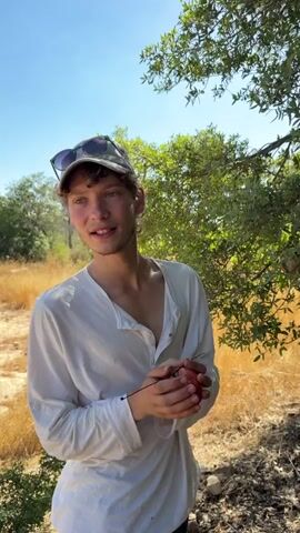 The video shows a young person wearing a white long-sleeved shirt and a white baseball cap with sunglasses on top. They are holding a small red fruit, possibly an apple or tomato, with a thin black stem attached. The person is standing outdoors in a dry, grassy area with trees and foliage in the background, under a clear blue sky. The lighting suggests it is daytime. The person appears to be looking off-camera to their left, then slightly shifts their gaze and adjusts their cap. They briefly open their mouth as if to speak. The video is shot from a slightly low angle, focusing on the person.