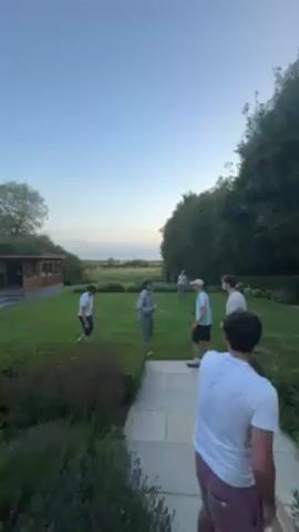 The video shows six young men on a grassy lawn. One man is standing on a stone walkway facing away from the camera, holding a cricket bat. He appears to be watching the other five, who are on the lawn. The group is interacting, with the man holding the bat seemingly involved in a discussion or playful argument. The setting includes a building with a porch in the background, as well as a lush green landscape with trees and a view of a distant field under a twilight sky.