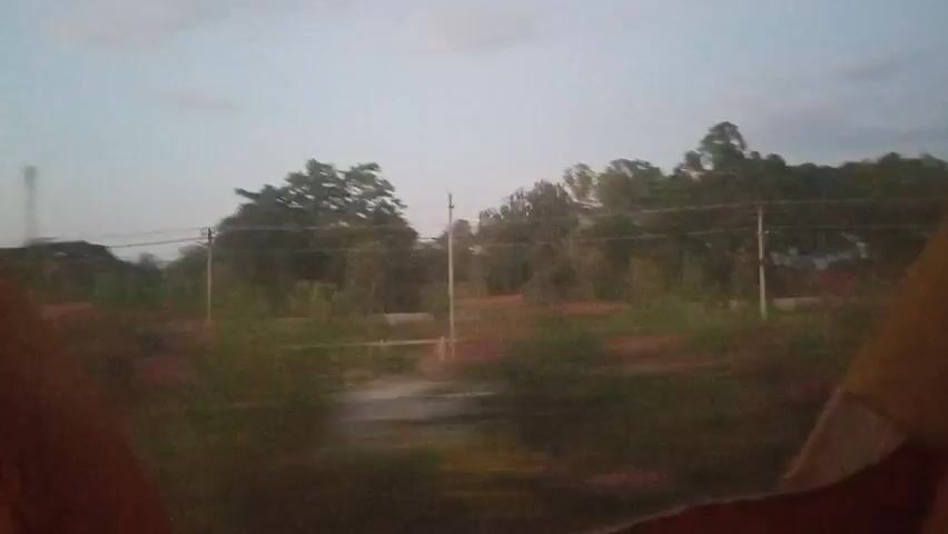 The video shows a blurry view of a landscape passing by, likely from a moving vehicle. The sky is overcast, and there are trees and vegetation visible. In the distance, there are some structures that appear to be telecommunication towers and possibly buildings. The motion blur suggests a high speed of movement, making it difficult to discern specific details of the environment or any potential occupants of the vehicle. The scene appears to be rural or semi-rural.