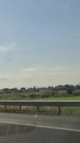 The video captures a view from inside a moving vehicle on a sunny day. The camera pans slightly, showing a roadside with a metal guardrail and a grassy field beyond. In the distance, a small village with houses and trees is visible under a bright blue sky with scattered clouds. The road ahead is visible, with other vehicles, including trucks, traveling in the same direction. The sun is high in the sky, casting a bright light.